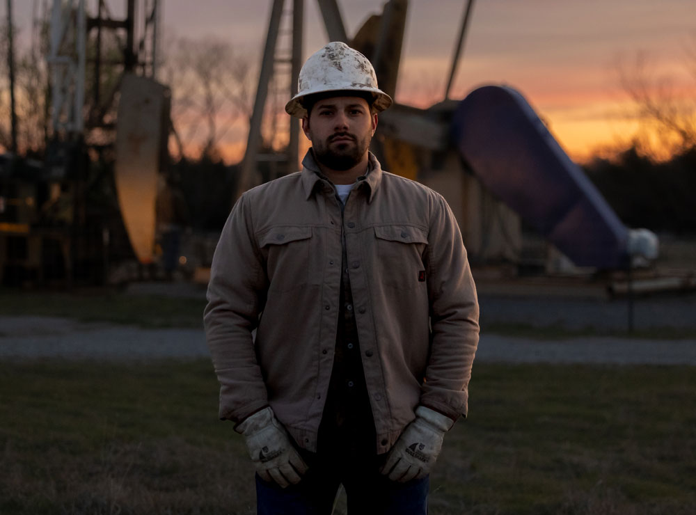 Un hombre, John Rodríguez, vestido con casco y guantes, posa con las manos frente a él, de pie frente a un gato de bomba de aceite.
